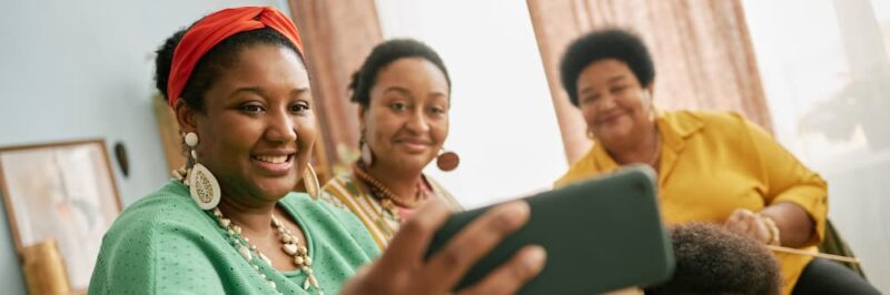 Three generations of Black women smiling together while looking at a smartphone to represent family health history and breast cancer awareness.